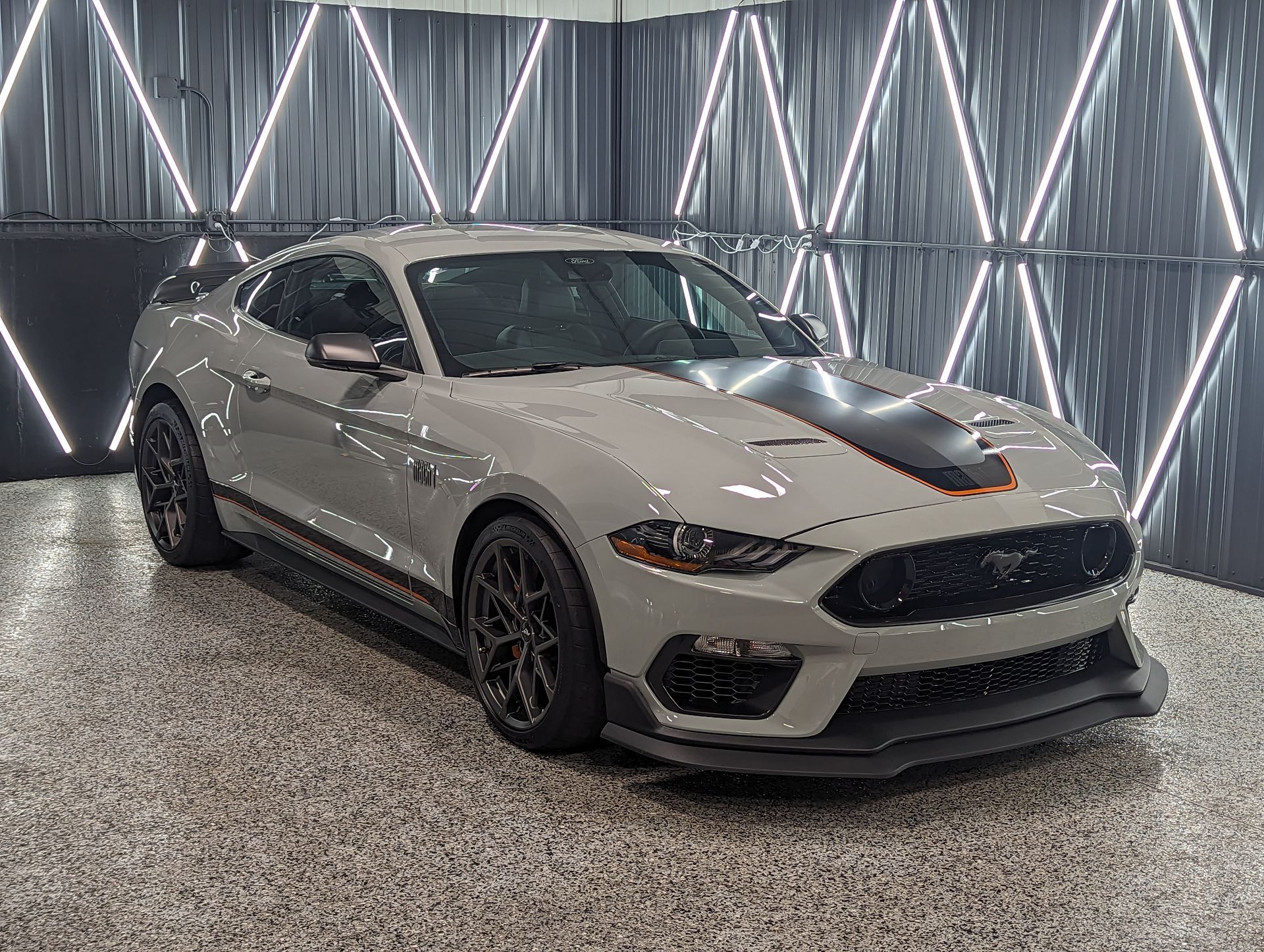 Gray Ford Mustang sports car with black racing stripes, inside a showroom.