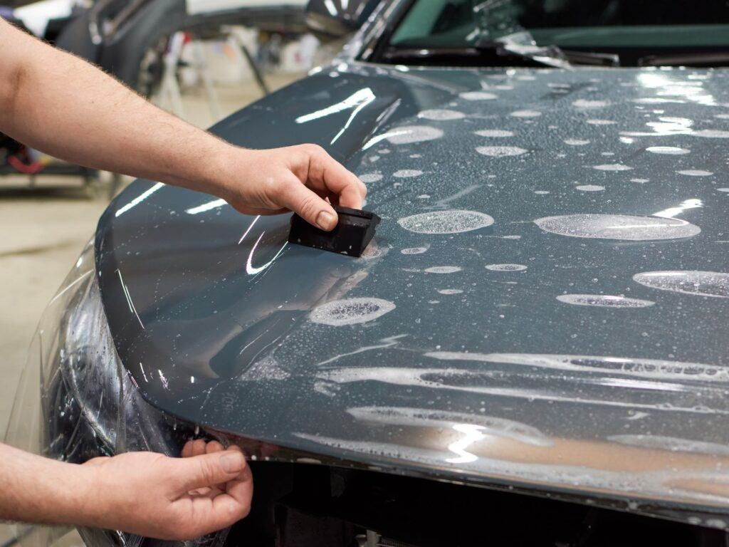 Hands applying clear protective film to a car hood, smoothing out bubbles with a tool.