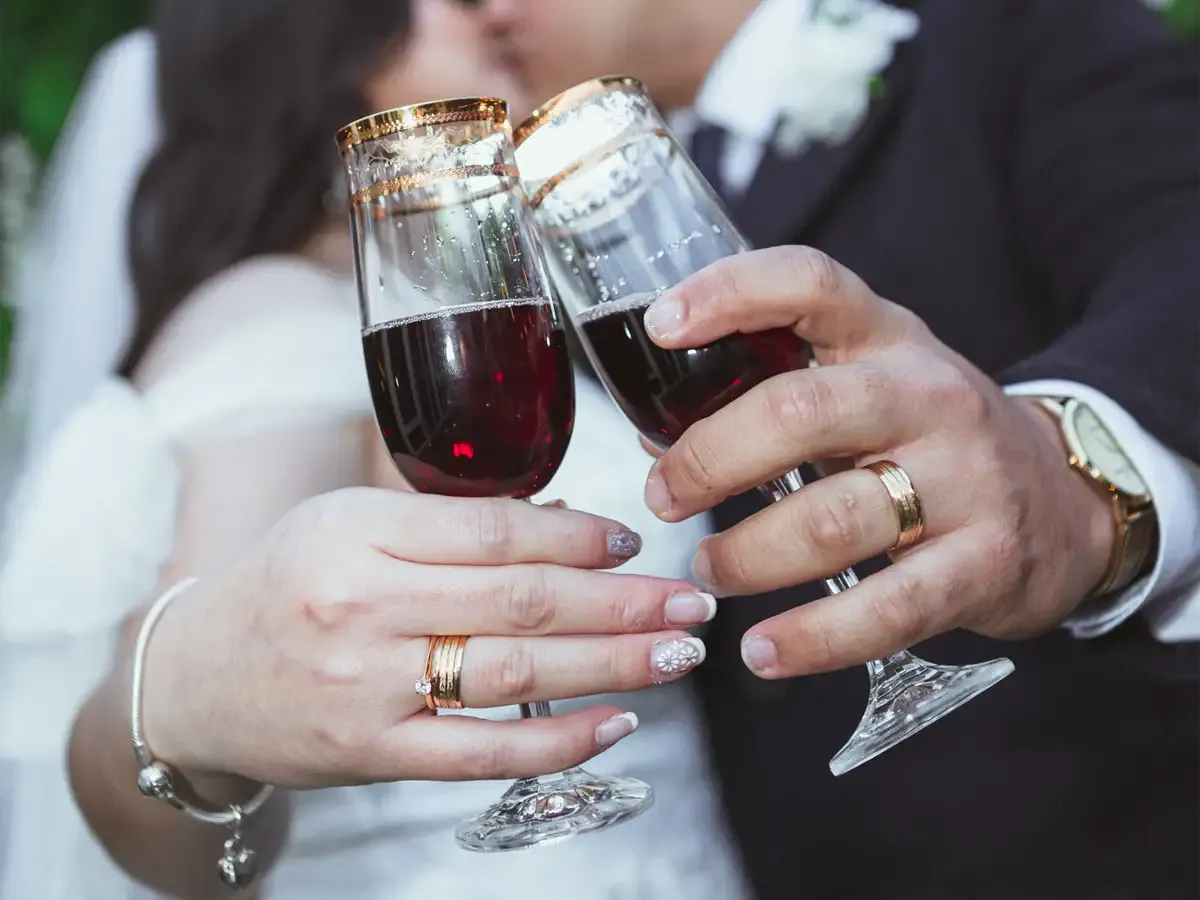 Couple clinking champagne flutes, wedding rings visible. Red liquid, golden rims, close-up, Bodas by Lina, Santa Clara, CA.