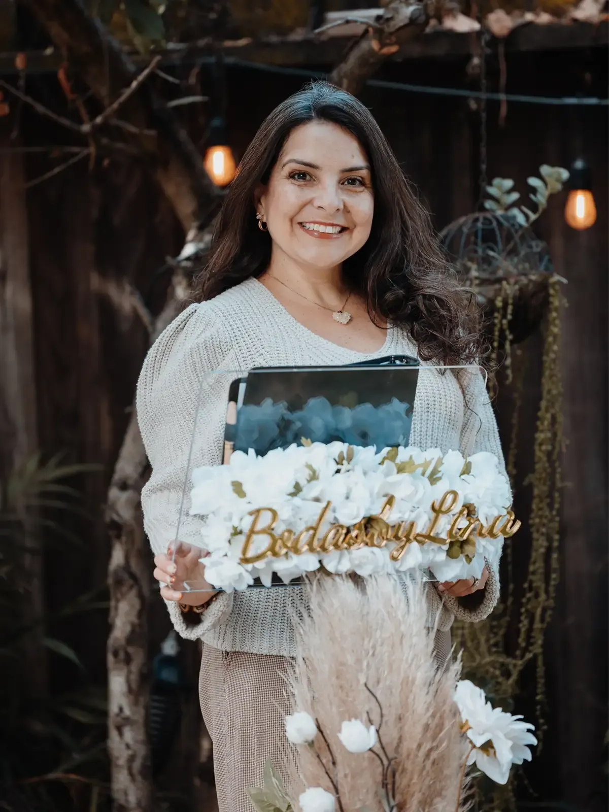 Lina of Bodas by Lina, Santa Clara, CA, Officiant, smiling, holding a sign in front of white flowers, in a garden setting.
