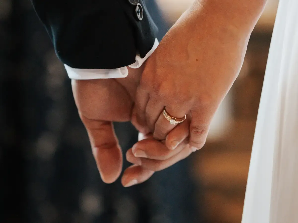 Hands clasped, one wearing a wedding ring, set against a dark suit and white dress, Bodas by Lina, Santa Clara, CA.
