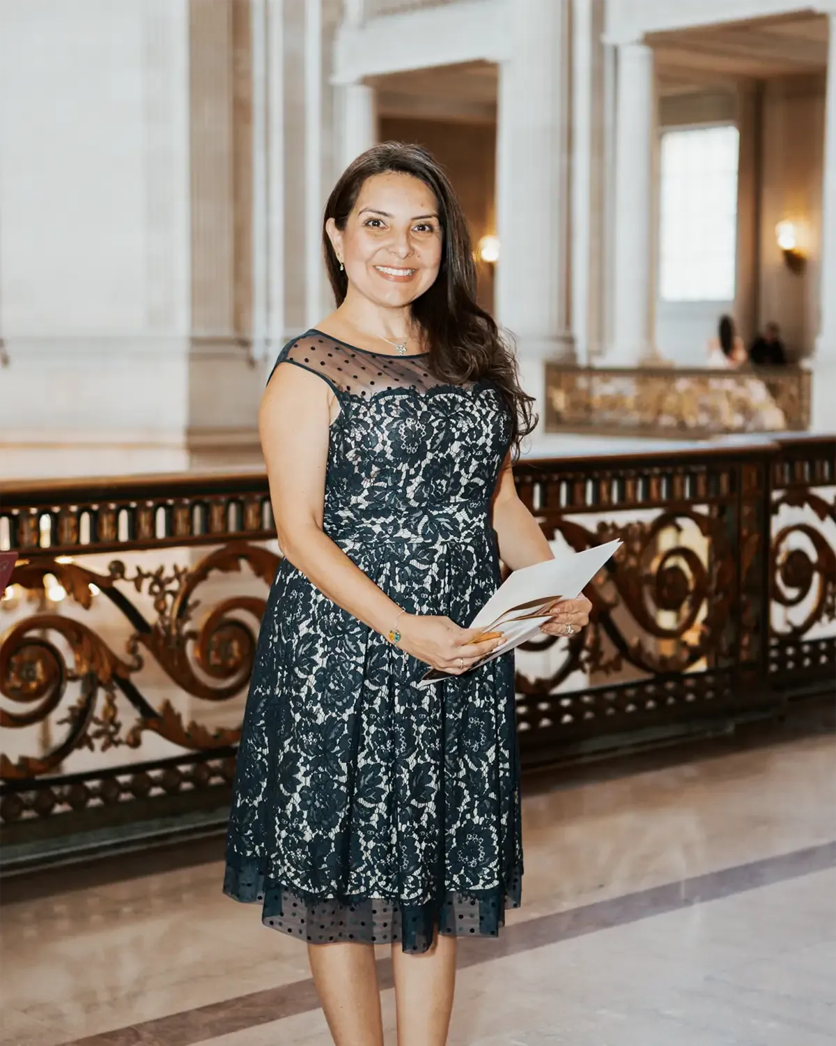 Lina of Bodas by Lina, Officiant, in Sant Clara, CA wearing a navy lace dress holds papers in ornate building, smiling.