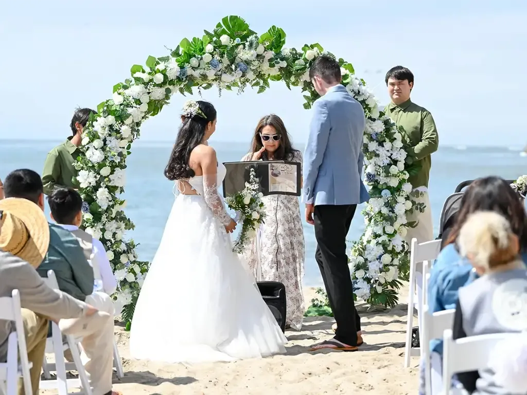 Wedding ceremony on the beach: Bride and groom under floral arch, officiant reading. Bodas by Lina, Santa Clara, CA.