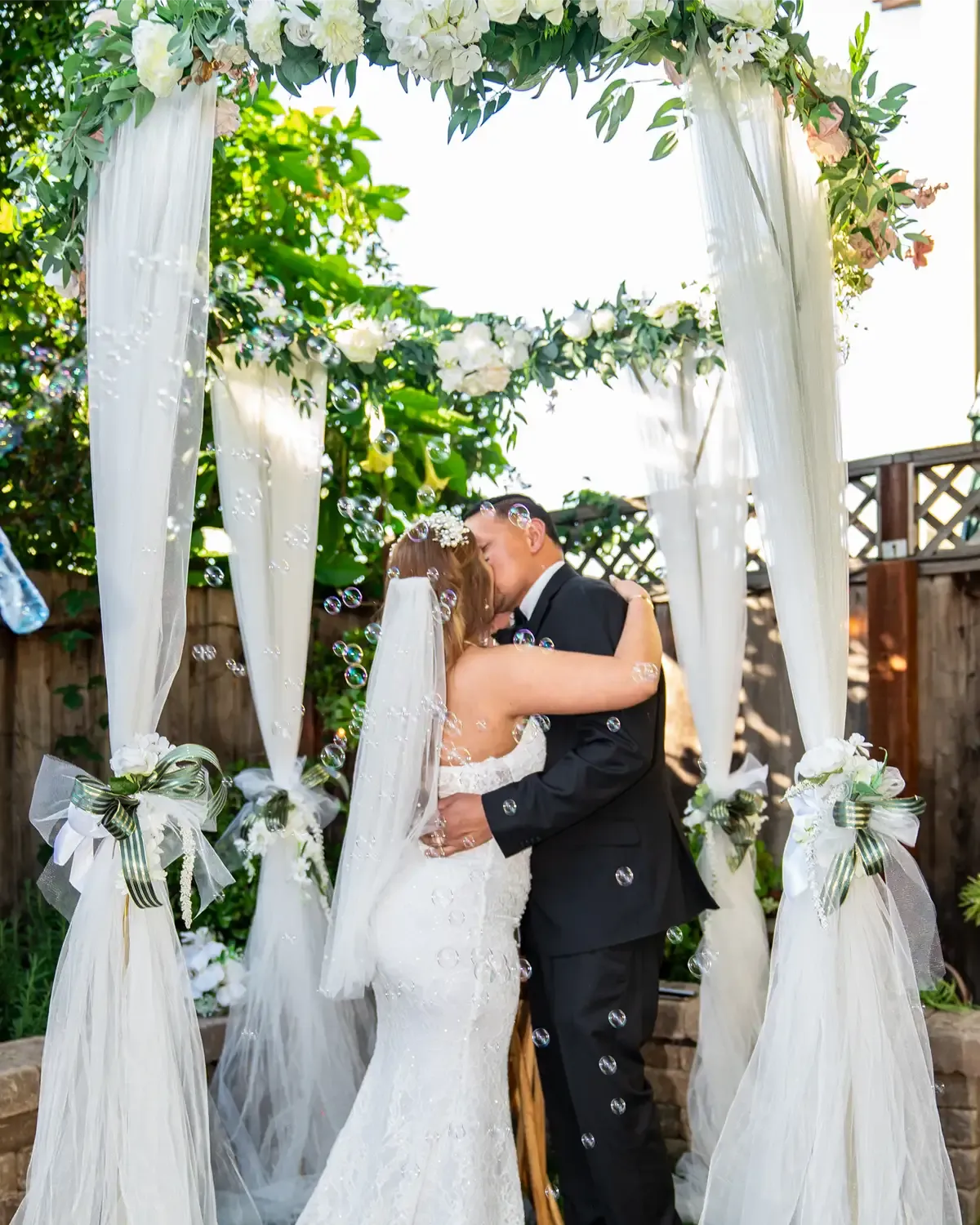 Bride and groom embrace, kissing under an arch decorated with flowers;  outdoor wedding, Bodas by Lina, Santa Clara, CA.