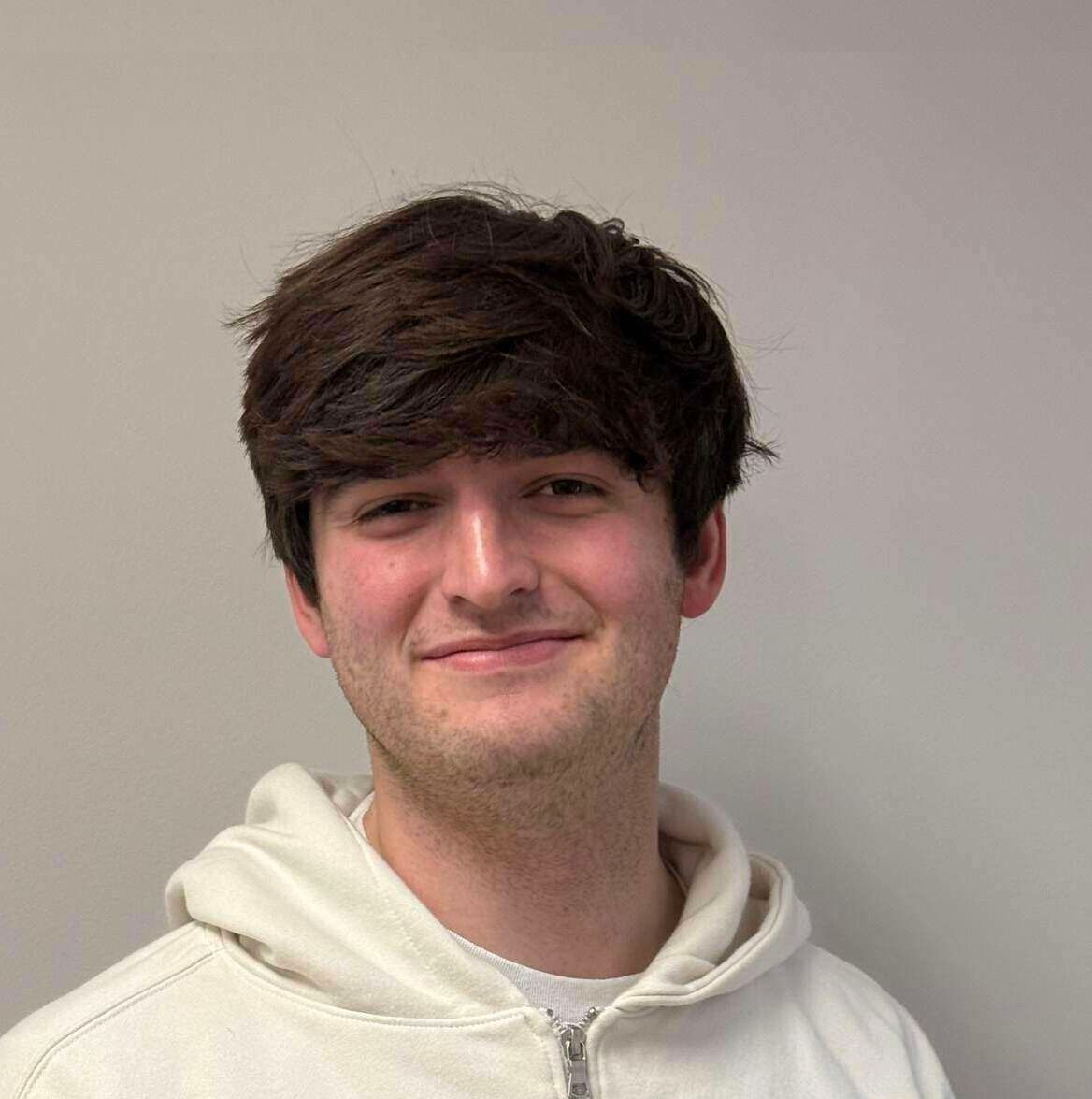 A young man with brown hair smiles while wearing a white zip-up hoodie. He is in front of a light gray wall.