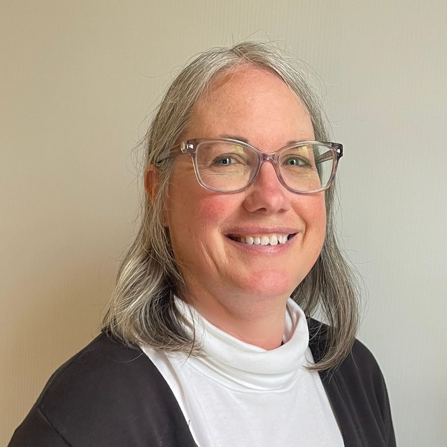 Woman with gray hair and glasses smiling, wearing a white turtleneck and black sweater.