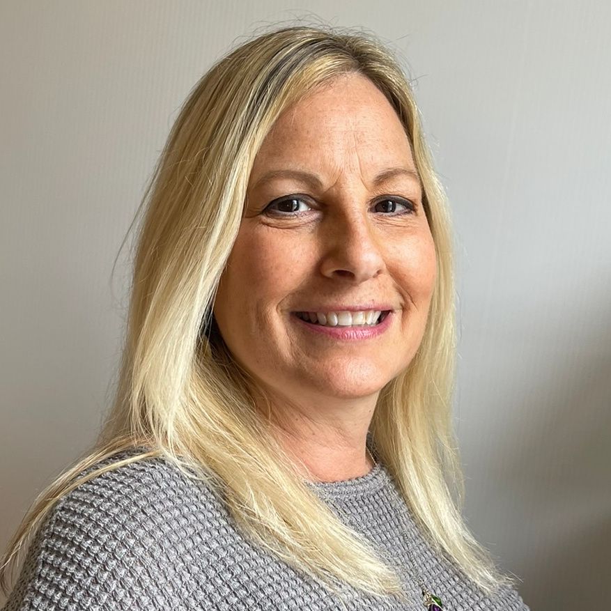 Blonde woman smiling, wearing a gray sweater, against a light-colored wall.