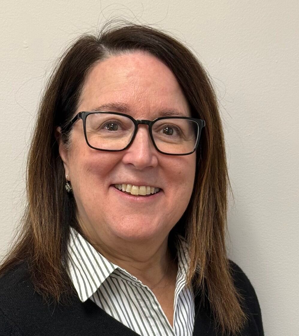 Woman with glasses smiling, wearing a striped shirt under a black sweater, against a neutral wall.