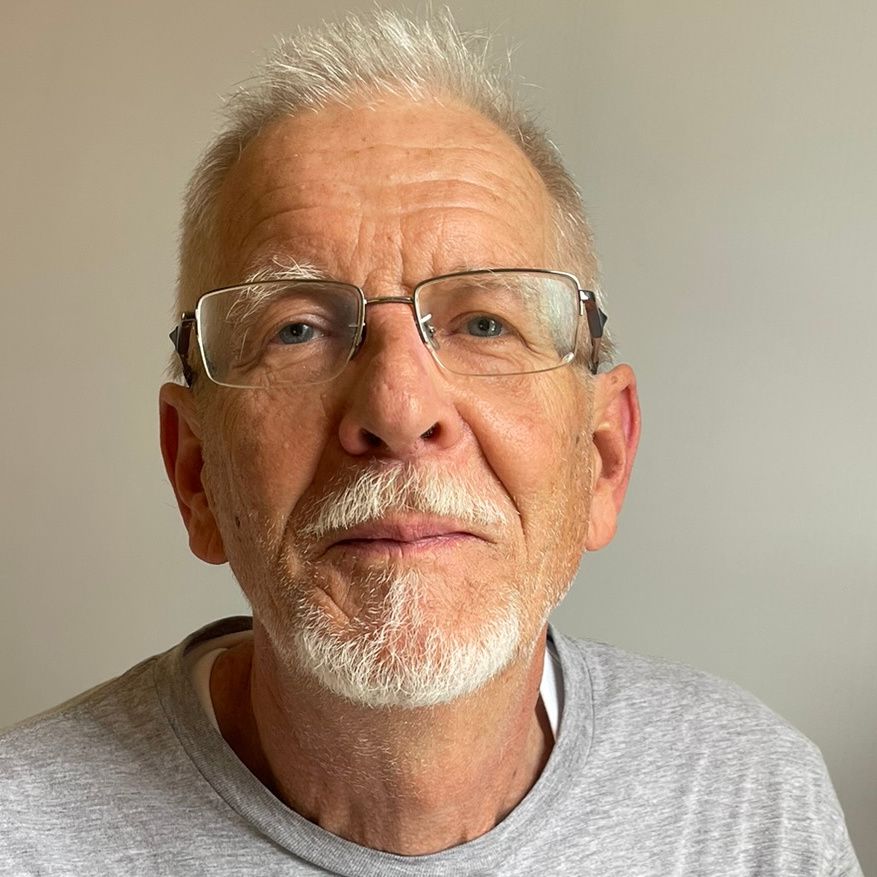 Older man with glasses, gray hair and beard, wearing a gray shirt, looking at the camera.