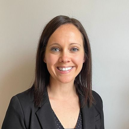 Woman with dark hair and a friendly smile wearing a black blazer, posing in front of a neutral wall.