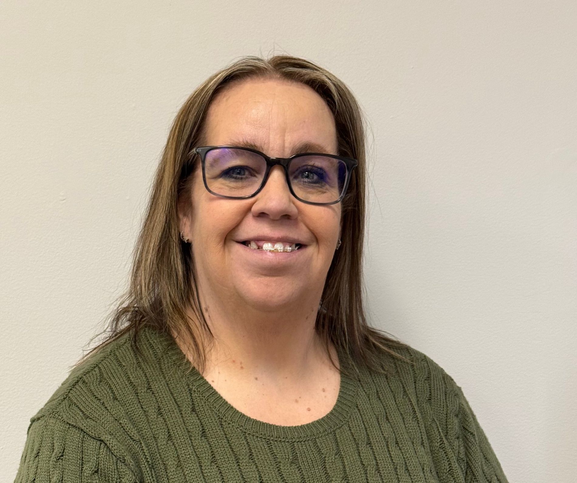 Woman with glasses smiling, wearing an olive green sweater, against a plain wall.