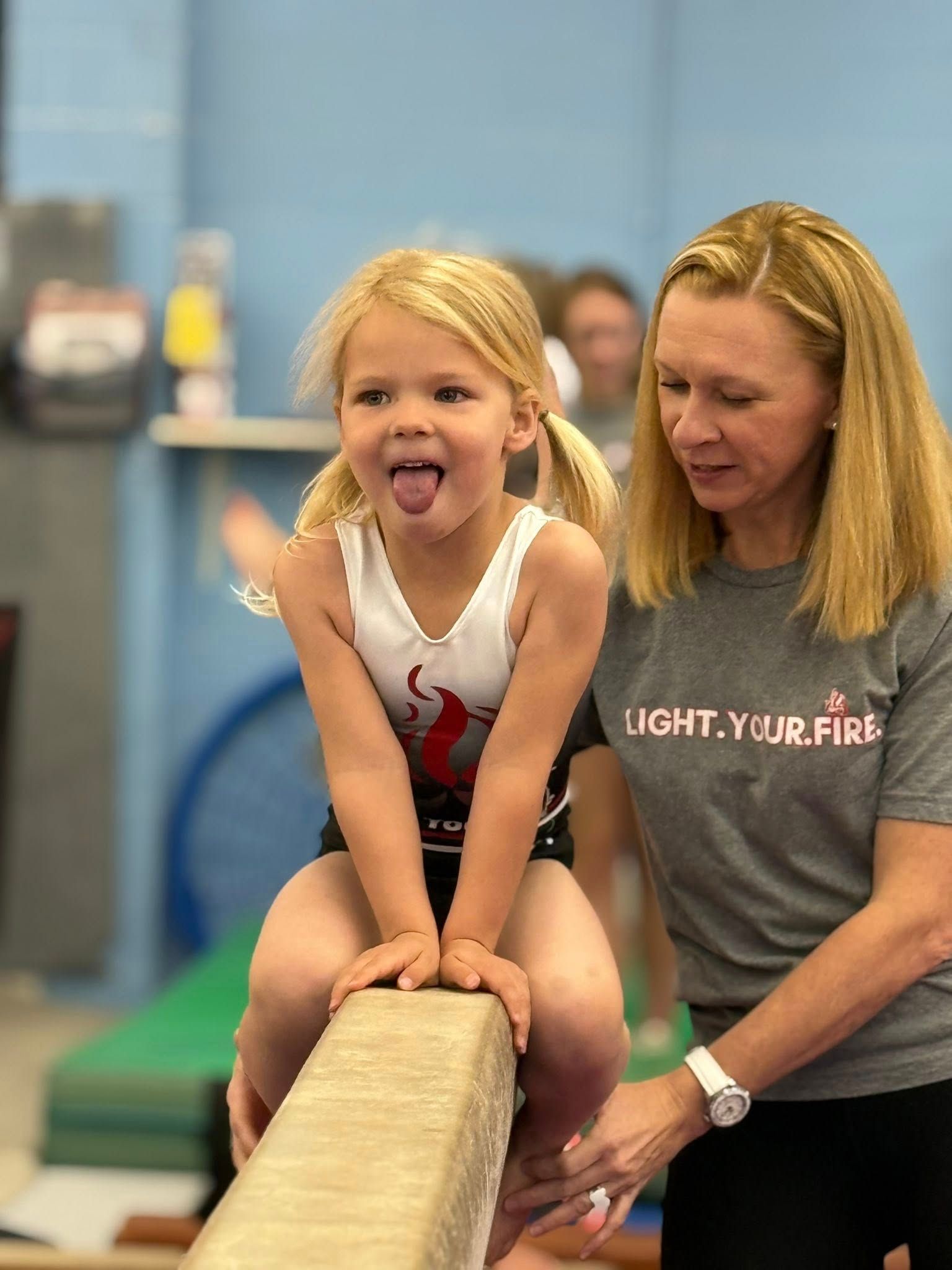 Hard at work processing your request! A young girl with pigtails sticks her tongue out while sitting on a balance beam, watched by a coach in a gym.