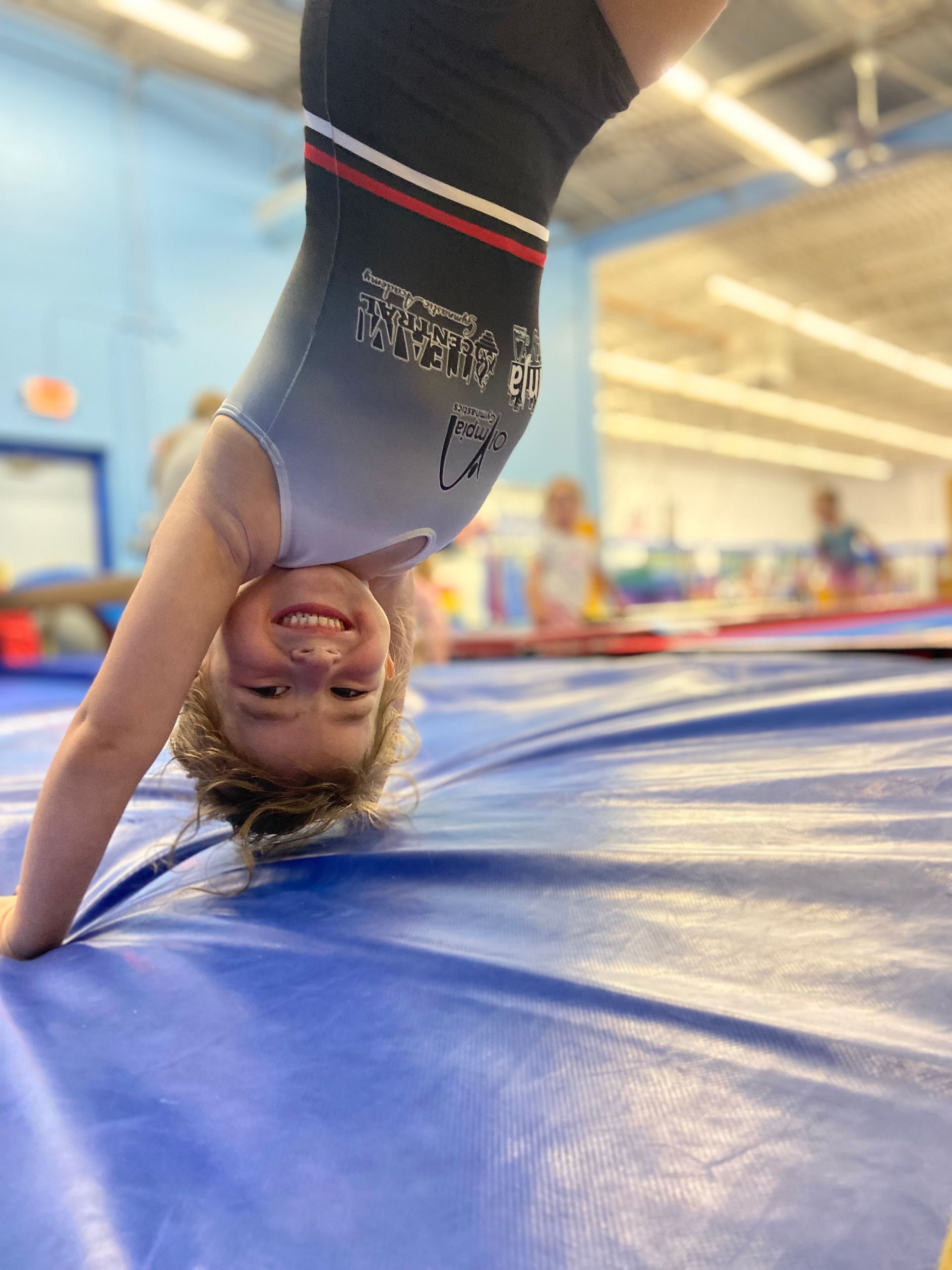Girl in pink leotard on a gymnastics bar, instructor assisting with leg stretch. Indoors, gym setting.