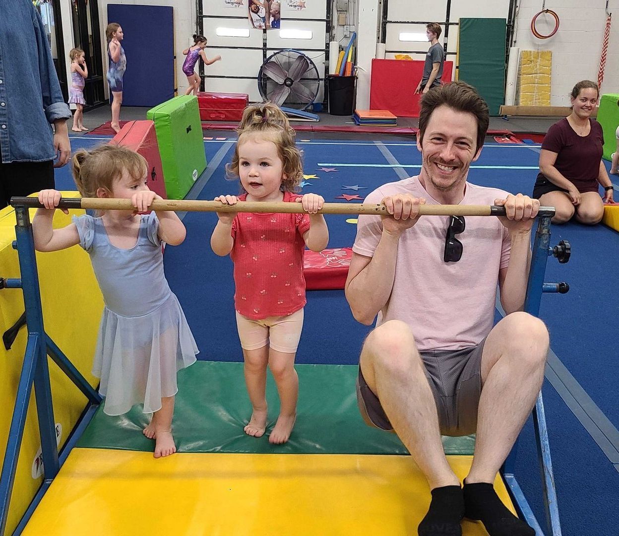 A man and two young girls hang from a horizontal bar in a gym. The man is smiling. Others are in the background.