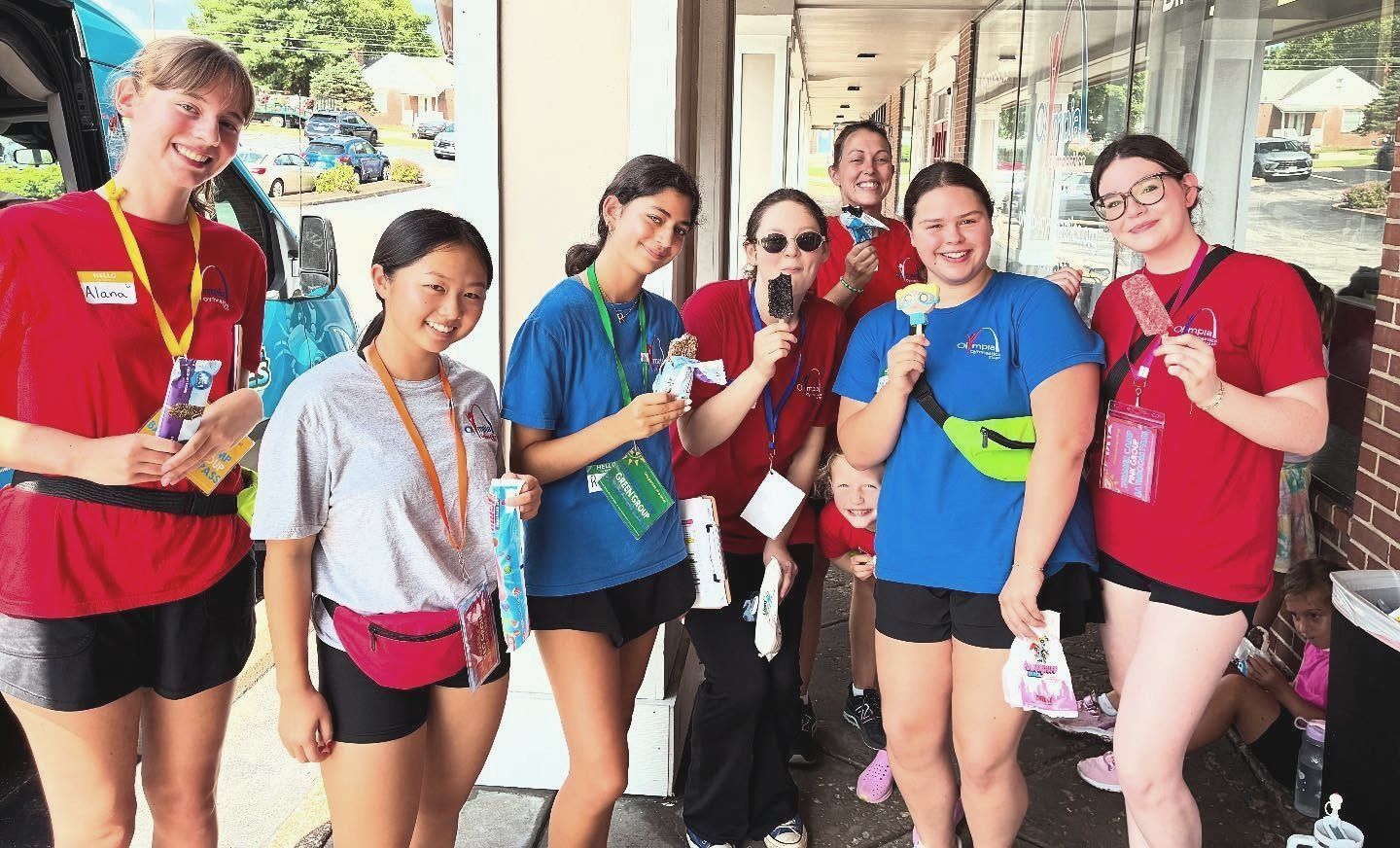 Group of young people smiling, wearing colored t-shirts and fanny packs, outdoors in front of a building.