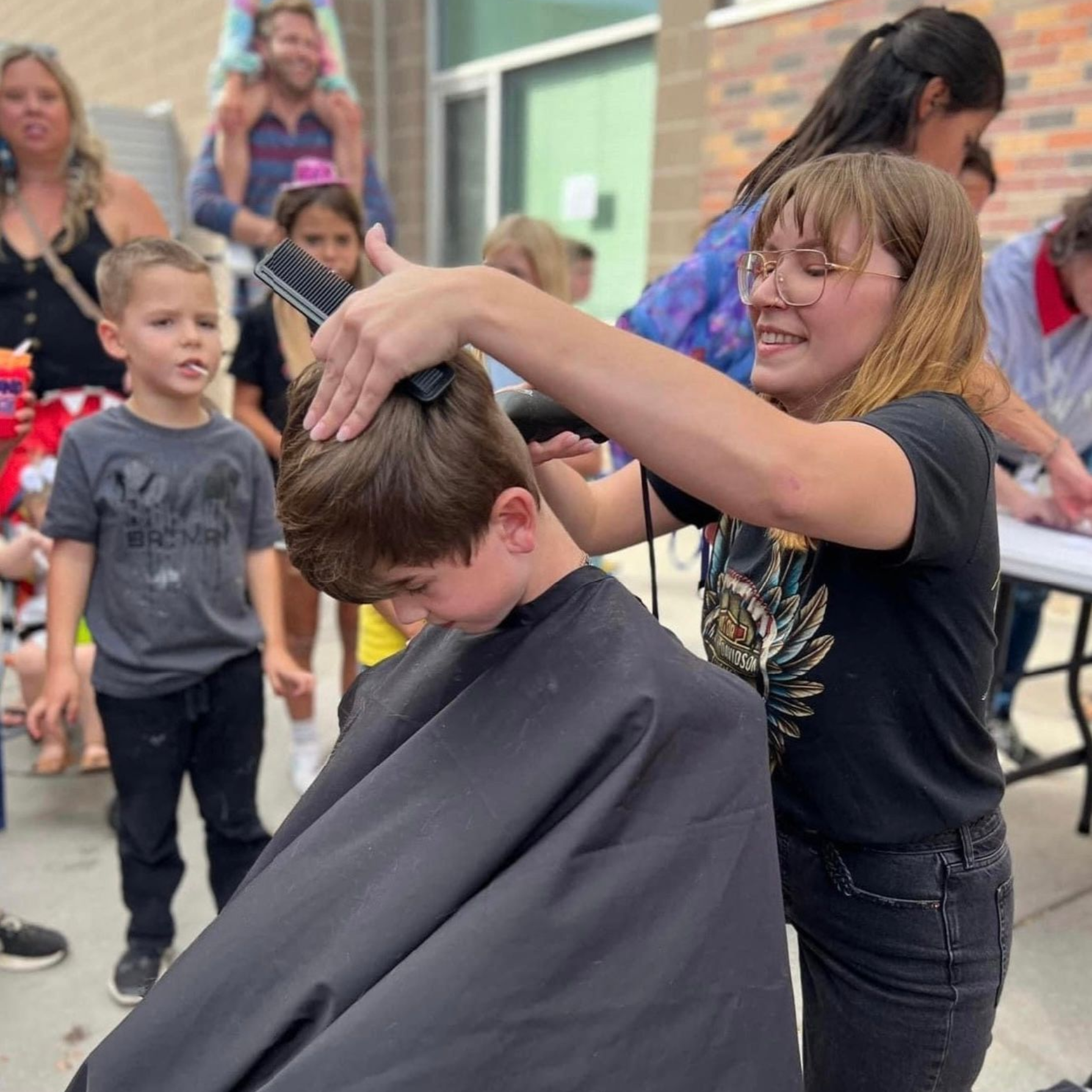 Woman giving a boy a haircut outdoors; other people watch.