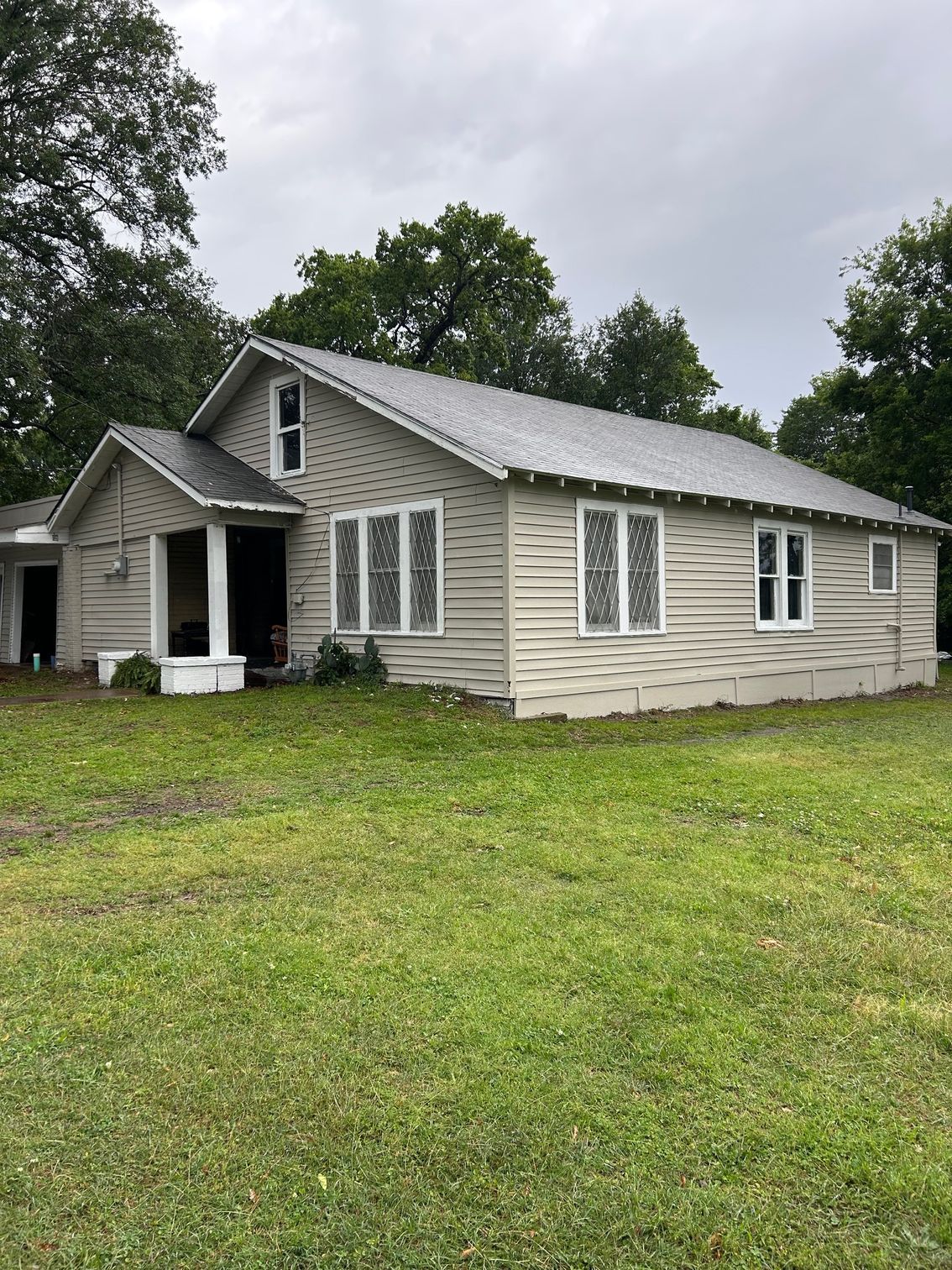 Small white house with a porch and gray roof, surrounded by a grassy yard and trees under cloudy skies