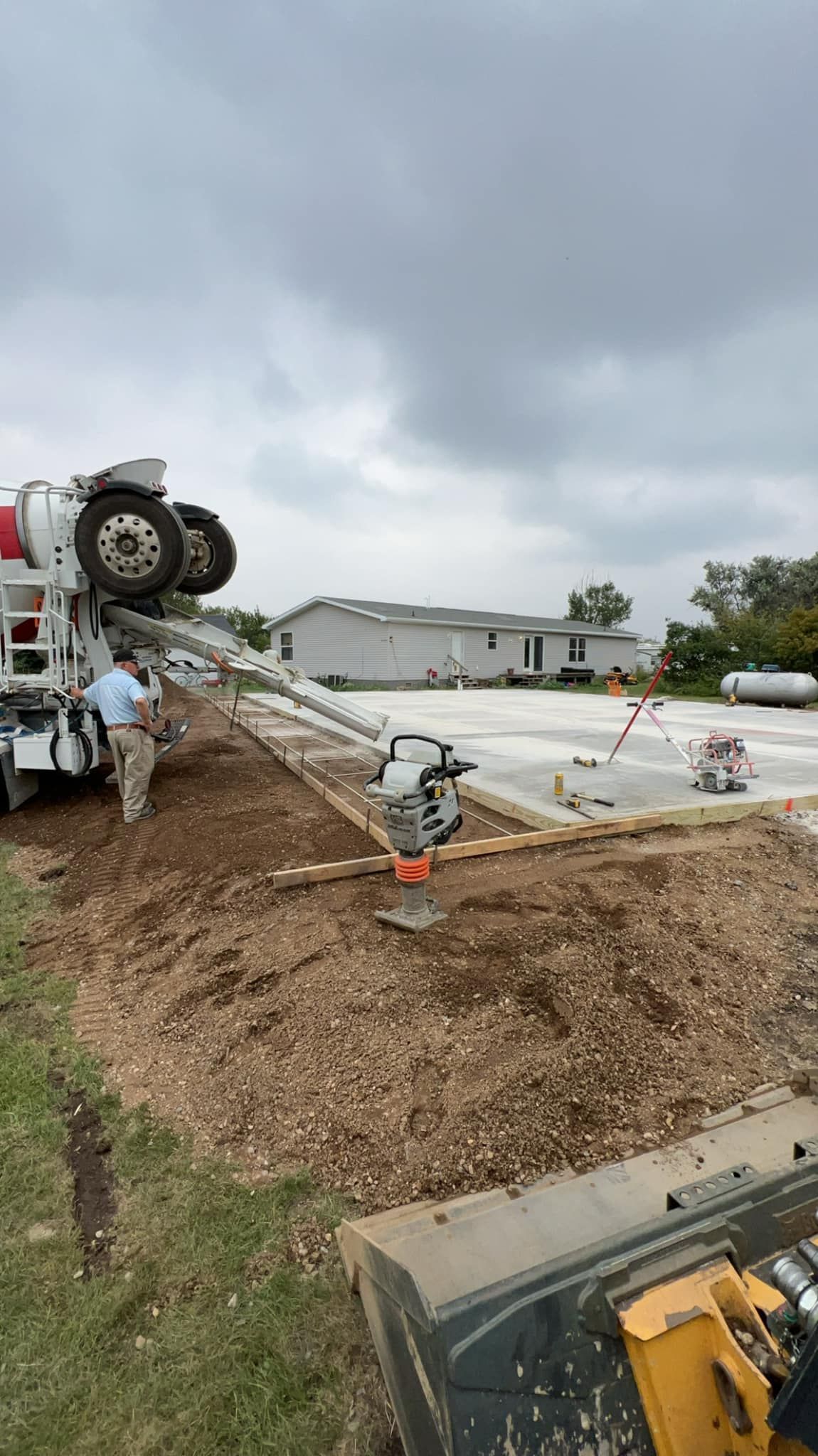 Concrete truck pouring cement into a prepared foundation; worker overseeing, overcast sky.