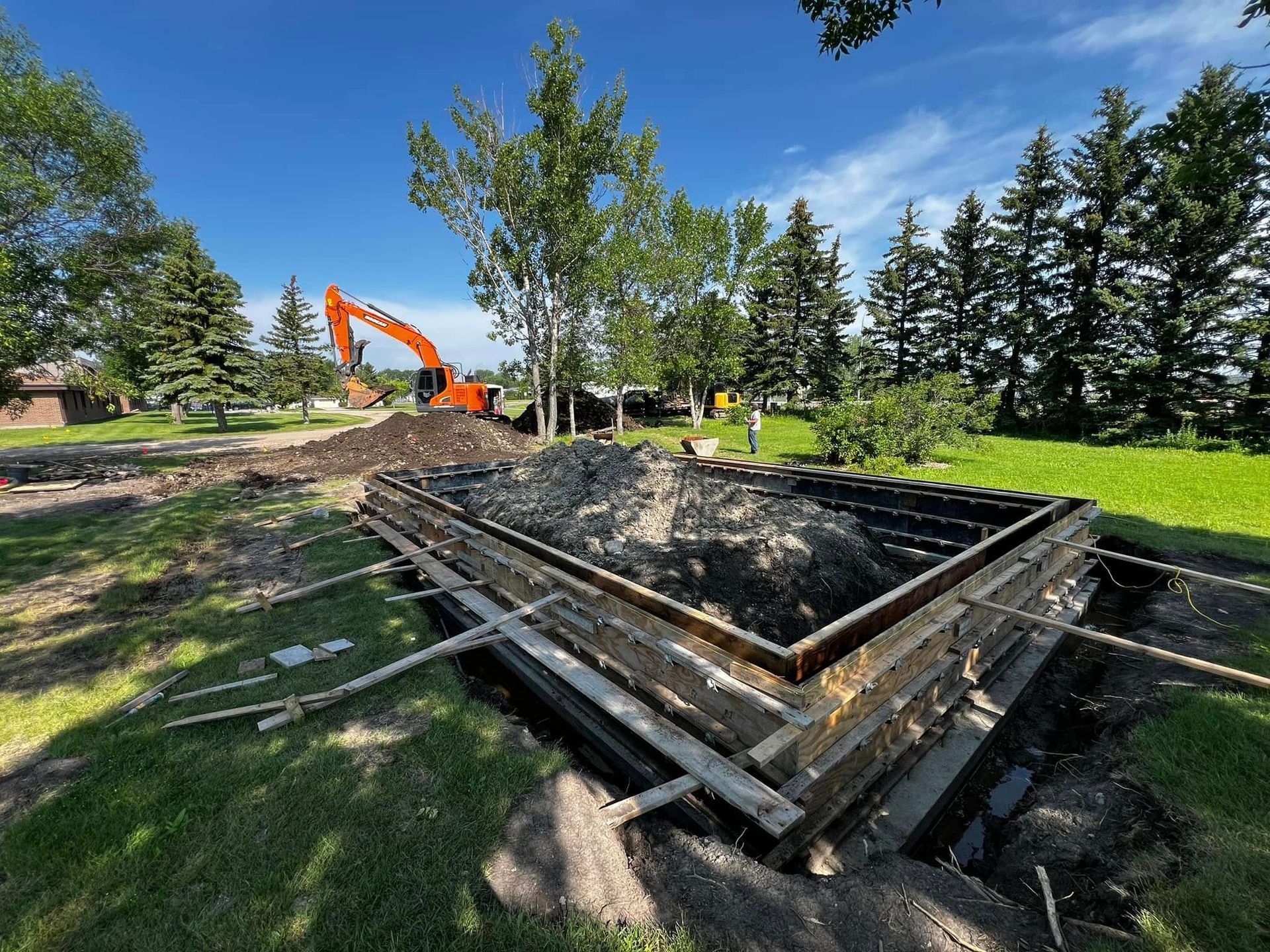 Excavation site with a rectangular concrete form. An excavator works in the background under a blue sky.