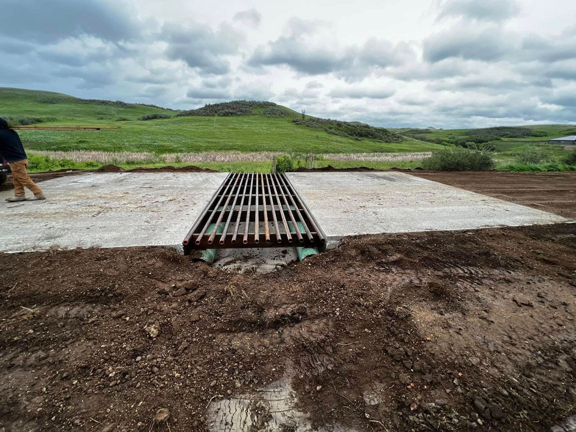Grate drain in gravel bed, earth surrounding. Man stands left. Green hills, cloudy sky.