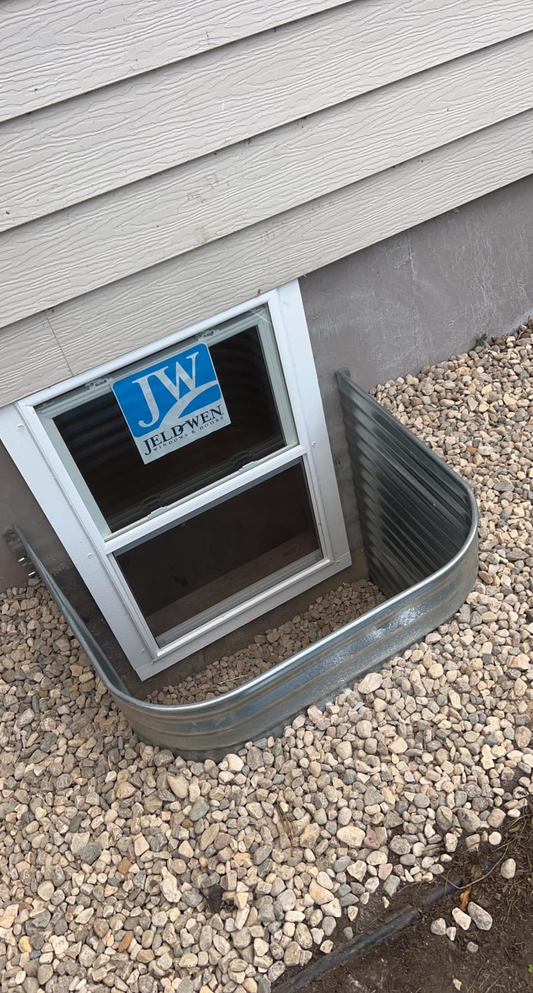 Window well installed below a home's siding. The window has JWZ branding. Gravel ground.