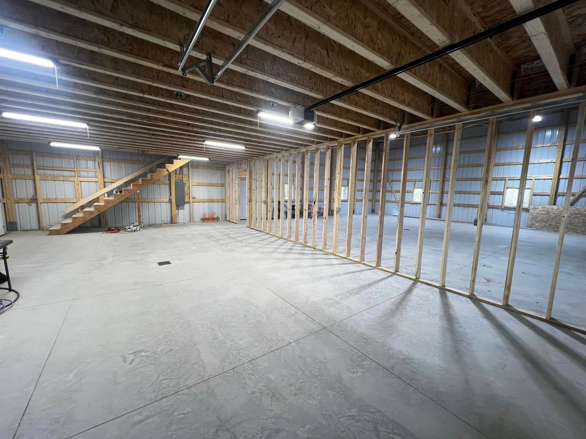 Interior of a large unfinished building with exposed wooden framing and concrete floor; stairs on the left.