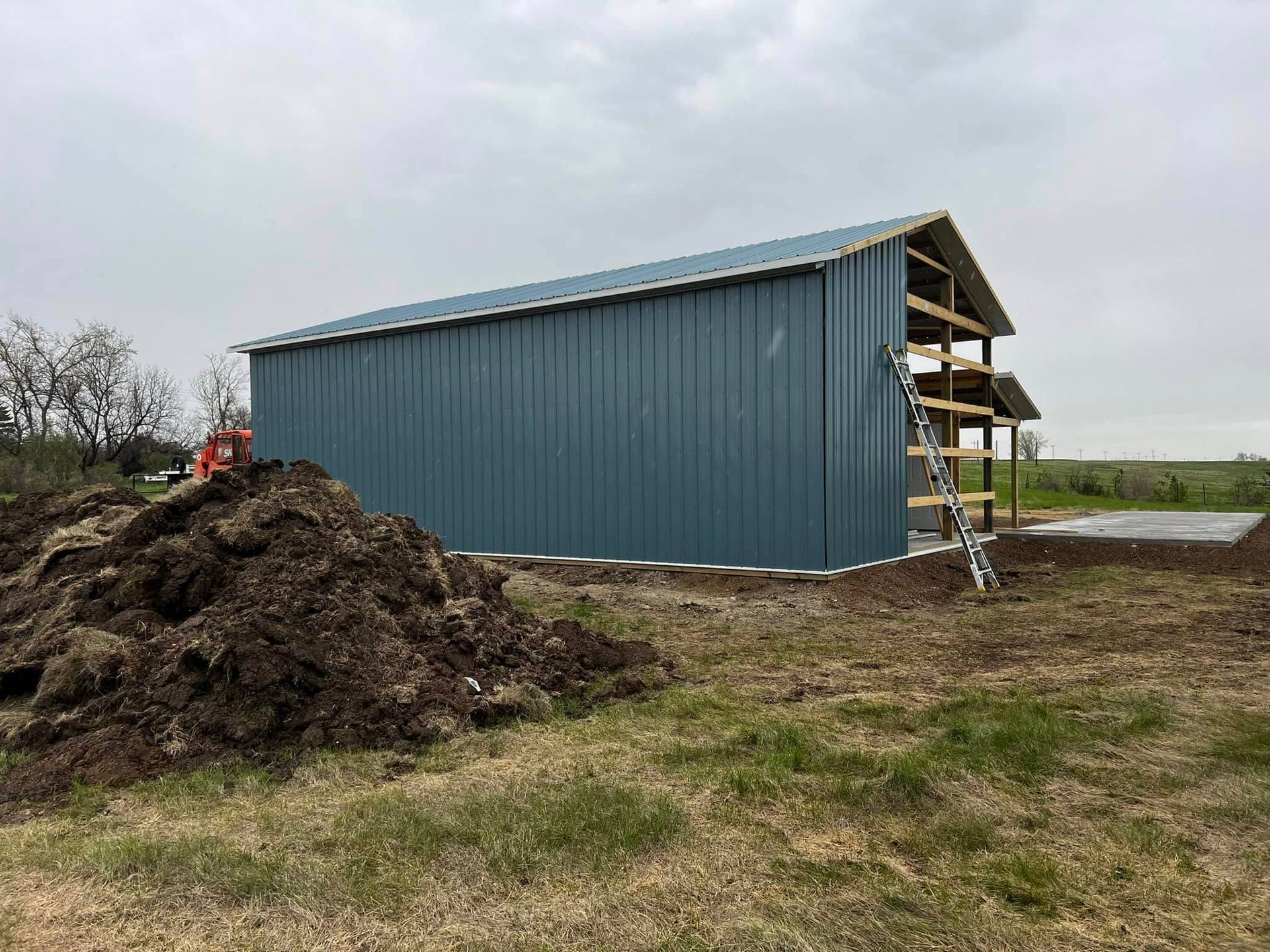 Blue metal building under construction on a grassy lot, with a pile of dirt in the foreground.
