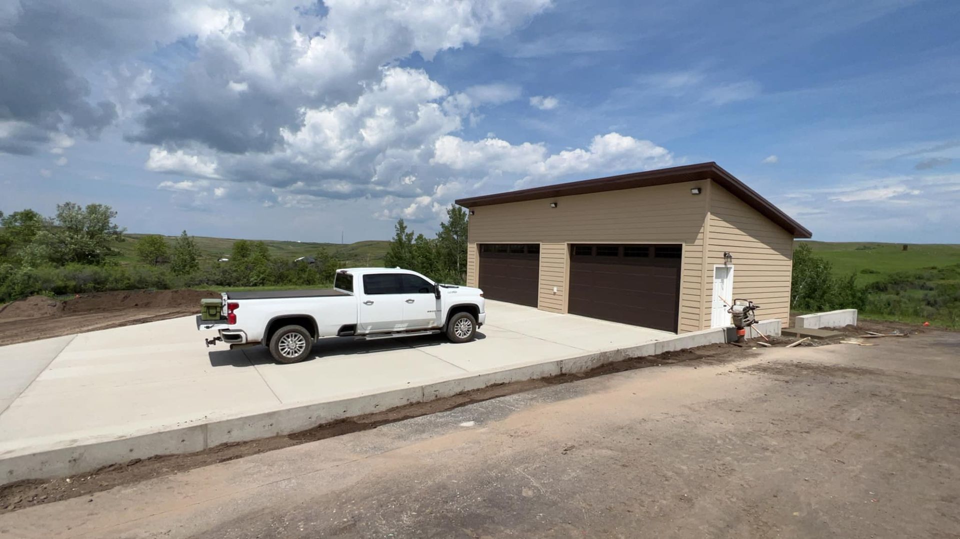 White truck parked on a concrete driveway in front of a tan garage with brown doors. Blue sky with clouds.