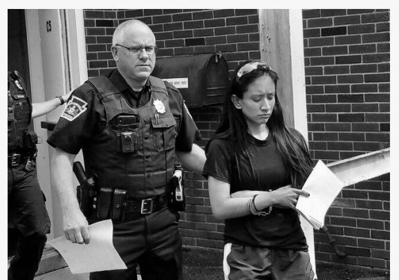 A police officer escorts a woman outside a building. The woman appears upset, holding papers.