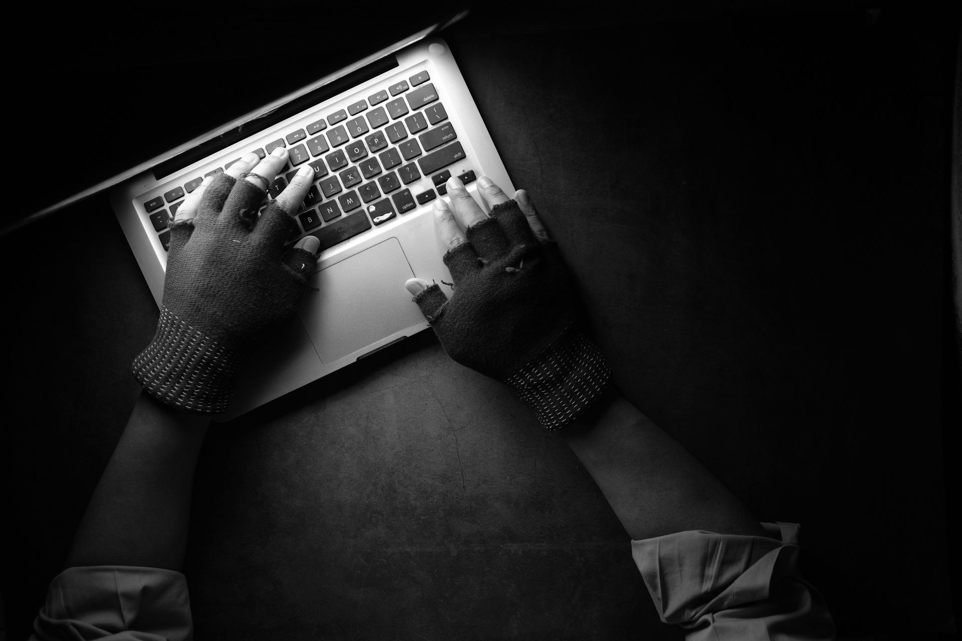 Hands in fingerless gloves typing on a laptop in a dimly lit setting.
