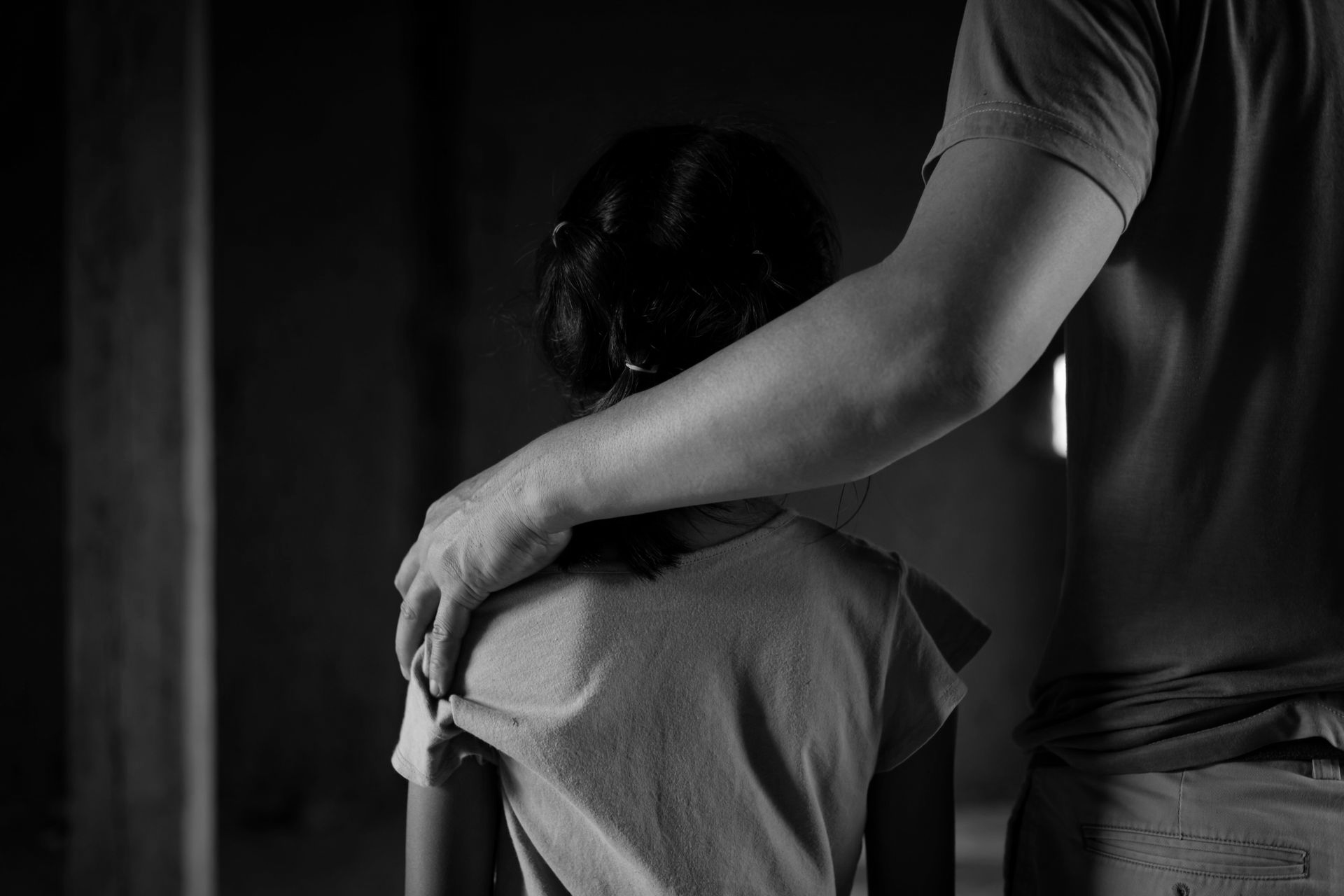 Black and white photo: Adult's arm around a child's shoulder, both facing away.
