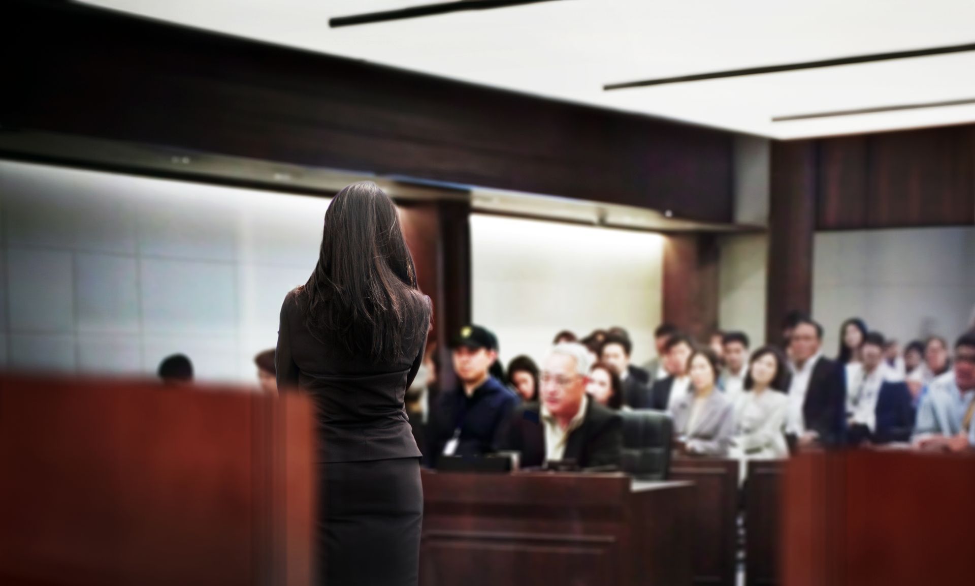 Woman in dark suit speaking in a courtroom, facing audience. Wooden paneling, bright overhead lighting.