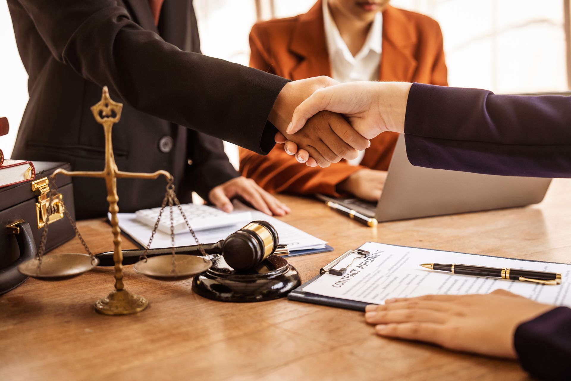People shaking hands at a law office; scales, gavel, legal documents on a wooden table.