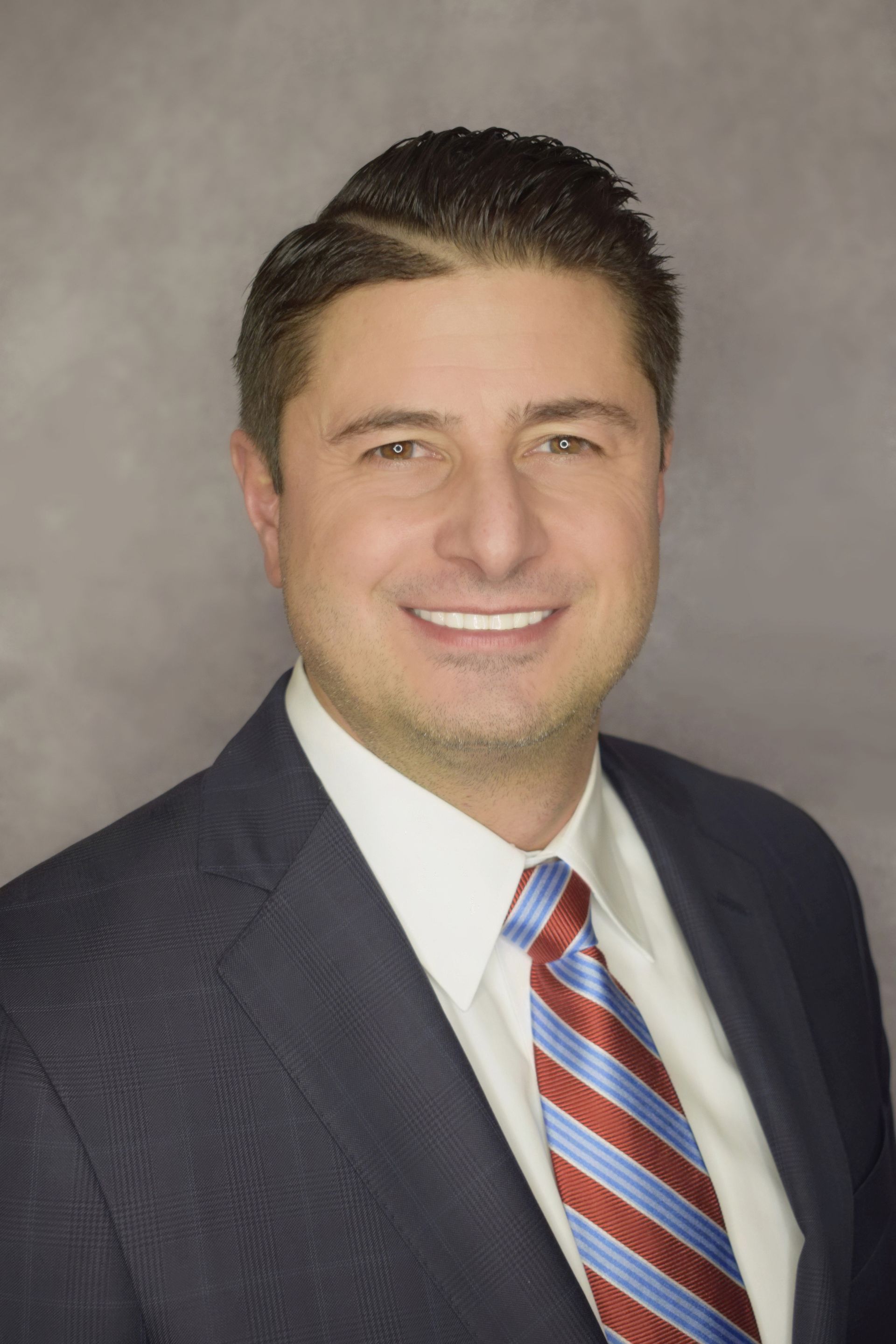 Man in navy suit, white shirt, and striped tie smiling at the camera. Gray background.