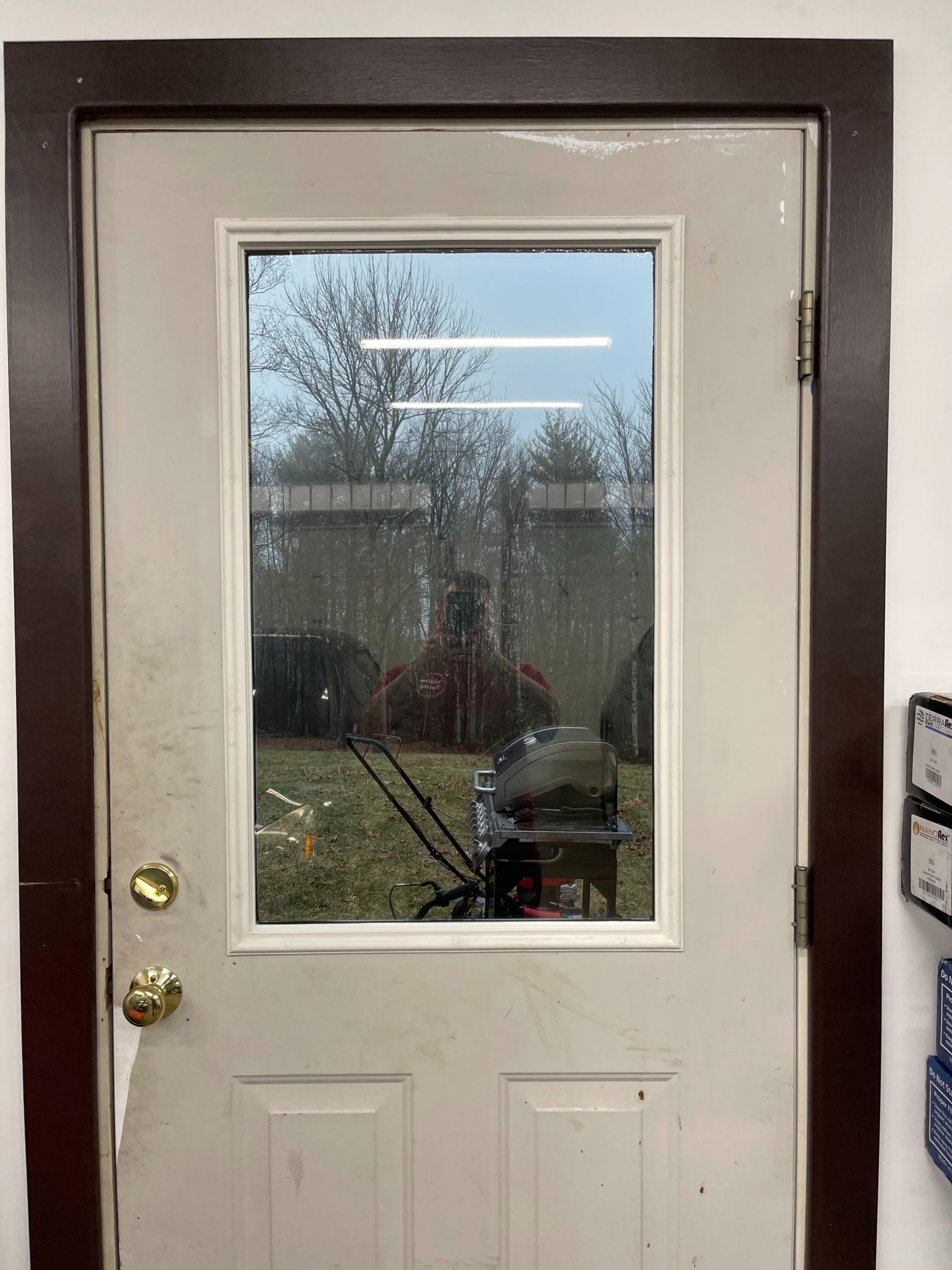 White door with a window; brown trim, gold doorknob, reflecting a yard and person.