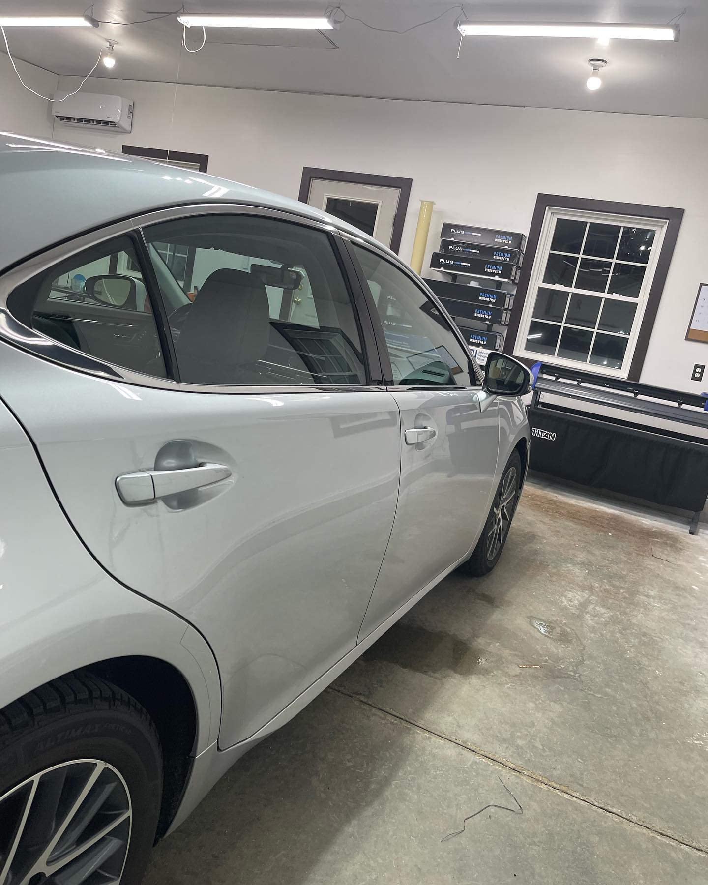 Silver car in a well-lit garage, side view. Tinted windows, visible tires, and garage interior.