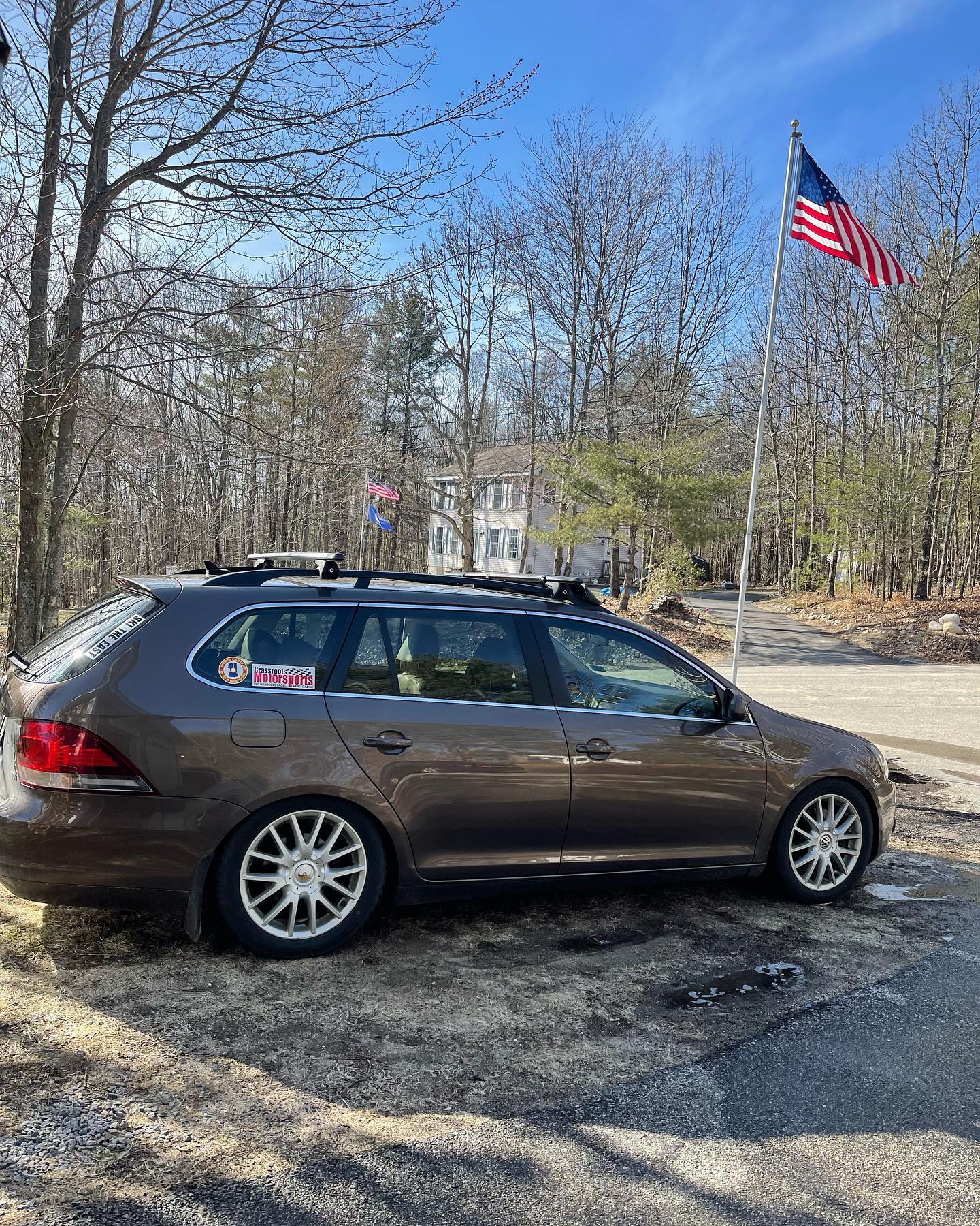 Brown station wagon with roof rack parked near an American flag, trees, and a clear blue sky.