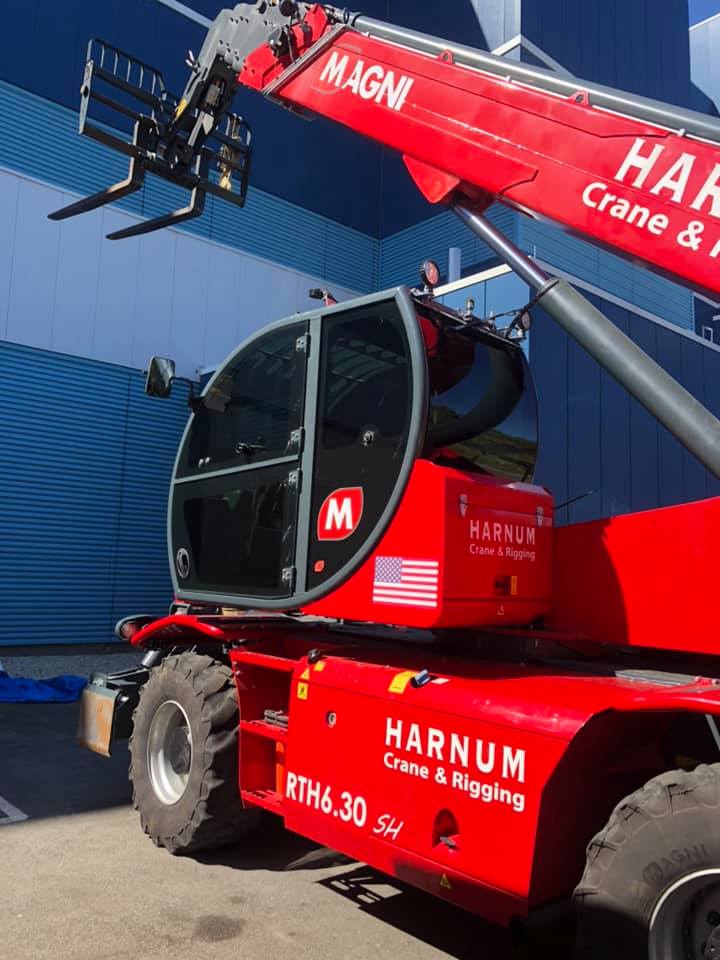 Red Harnum telehandler with forks extended, parked near a blue building.
