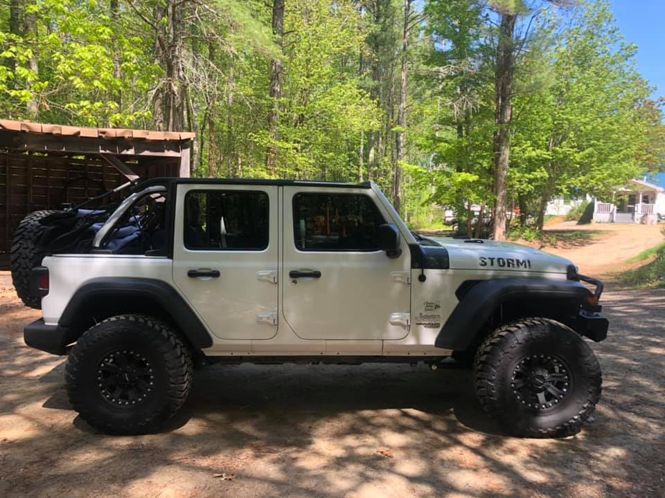 White Jeep Wrangler with large black tires parked on dirt road, trees in the background.