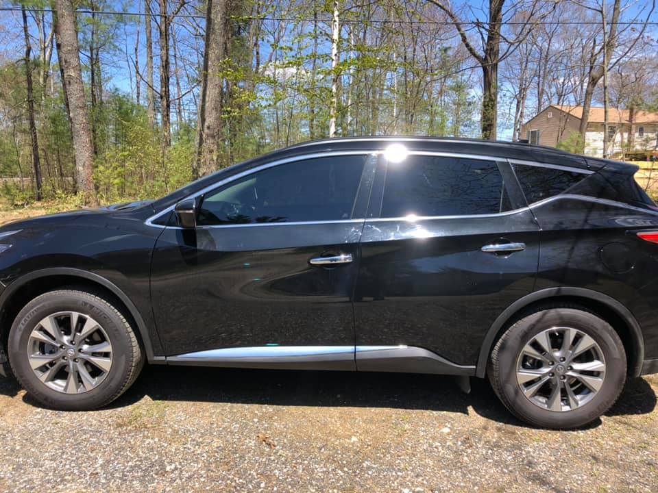Black SUV parked on gravel driveway with tinted windows, trees, and a house in the background.