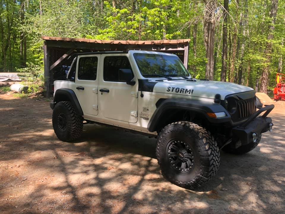 White Jeep Wrangler with black accents and large tires parked in a wooded area. 