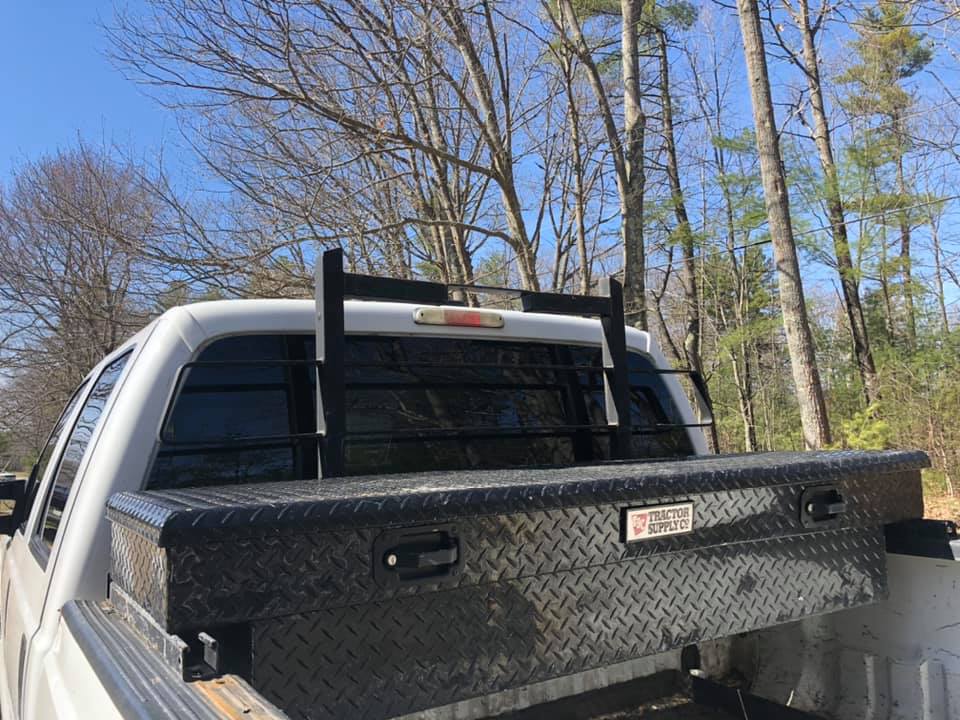 White pickup truck bed with black tool box, rack, and rear window guard against a tree-lined backdrop.