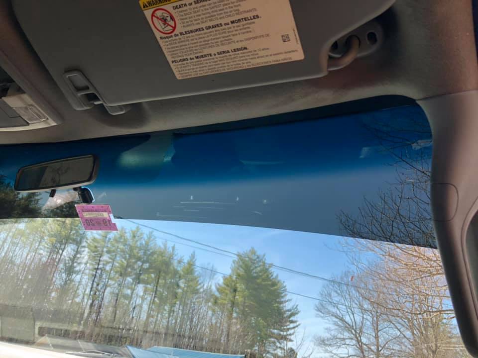 Interior view of a car looking out through the windshield at trees and sky; sun visor and rearview mirror visible.