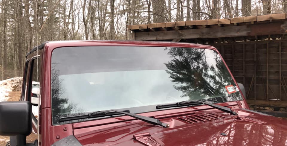 Red Jeep's windshield with a tinted strip at the top, parked near a wooden structure and trees.
