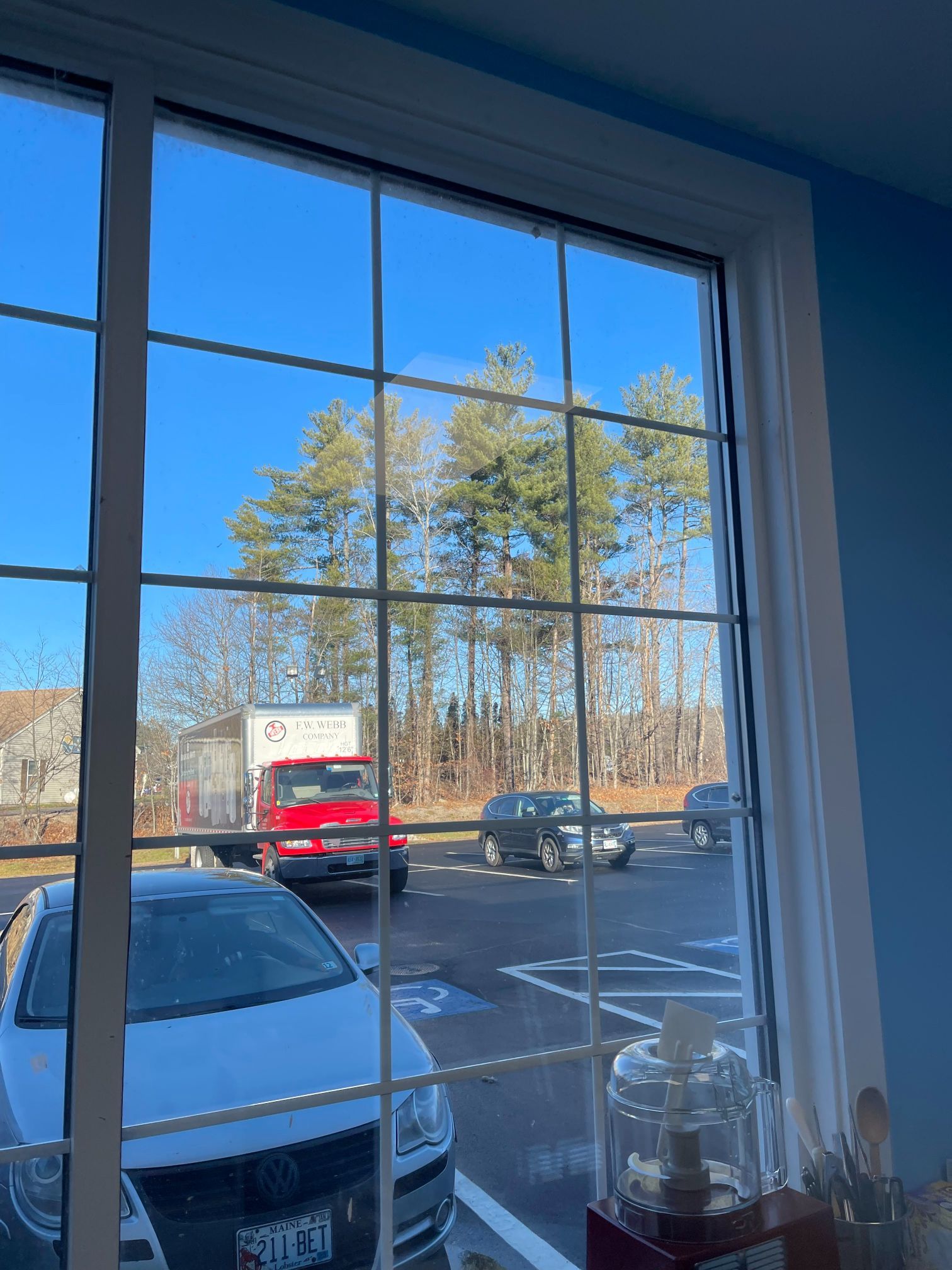 View through a window of a parking lot with cars, a red truck, and trees under a blue sky.