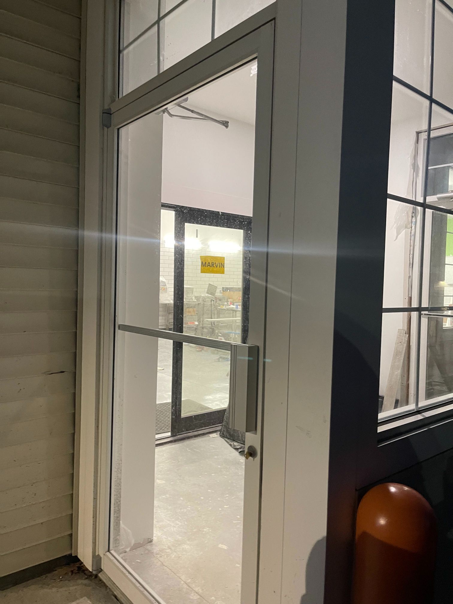 Glass door in a white frame, leading into a building. Adjacent window with grid. Orange bollard visible.