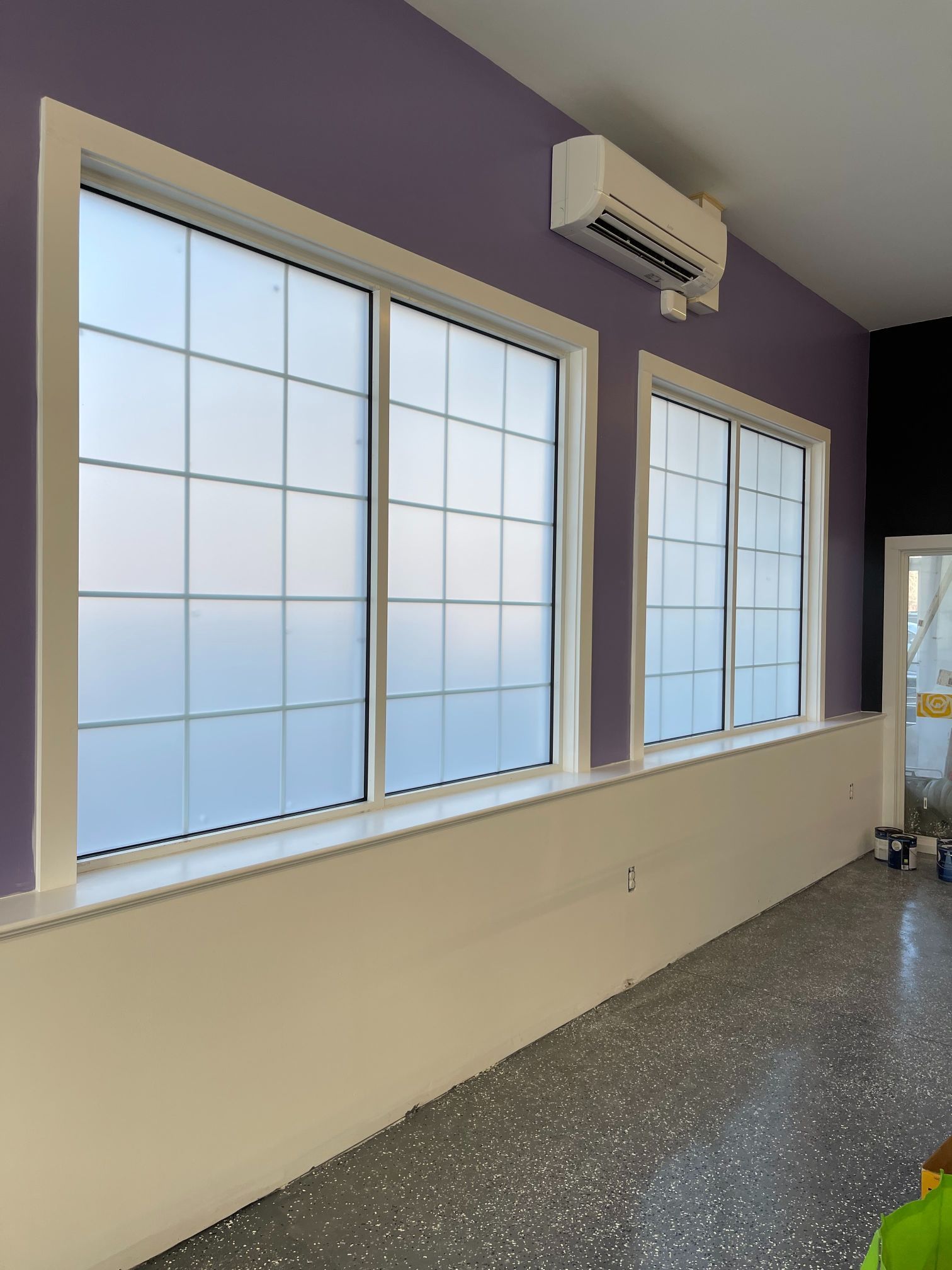 Two white-framed windows with frosted grids on a purple wall. White trim and baseboard, gray floor, and AC unit.