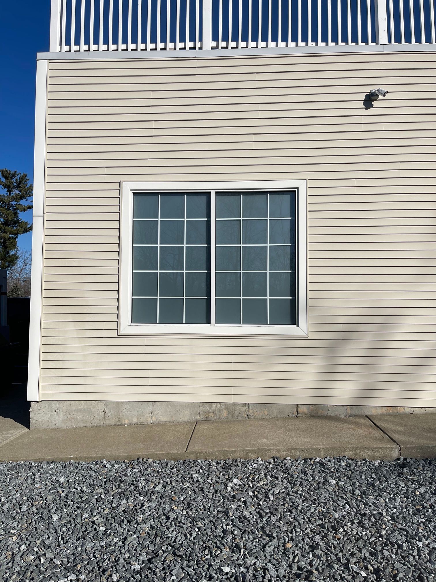 Exterior of a building with a white-framed window. Light tan siding, gravel ground, and a balcony above.