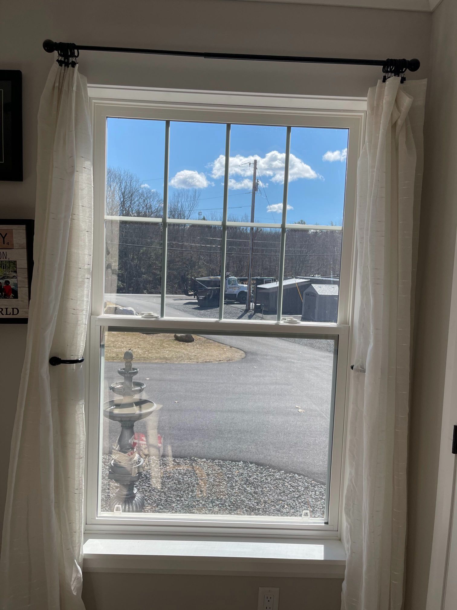 Window with white curtains, showing a bright blue sky with clouds and a view of a street.