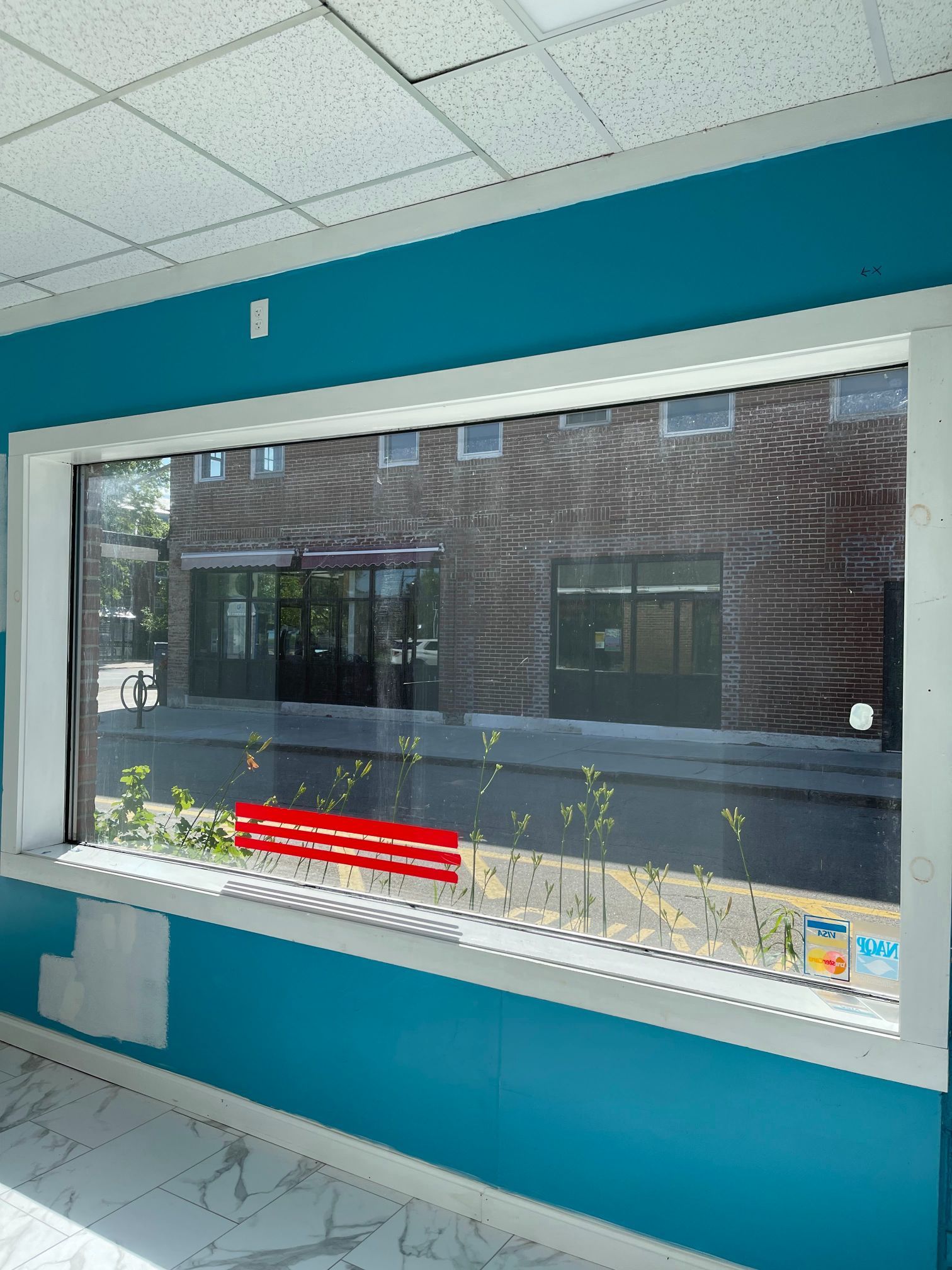 View of a building exterior through a window with white trim, turquoise walls. A red bench is visible outside.